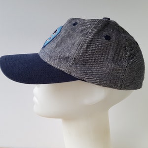 May include: A baseball cap with a navy blue brim and a gray speckled crown. The cap features a small patch on the side and a navy blue button on top. The cap is displayed on a white mannequin head.
