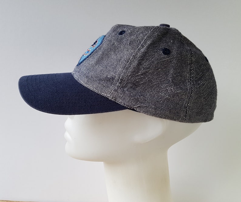 May include: A baseball cap with a navy blue brim and a gray speckled crown. The cap features a small patch on the side and a navy blue button on top. The cap is displayed on a white mannequin head.