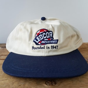 May include: A cream-colored baseball cap with a navy blue brim. The cap features the embroidered text "LEDCOR INDUSTRIES" above "Founded in 1947" in red, white, and blue. The cap is sitting on a wooden surface.