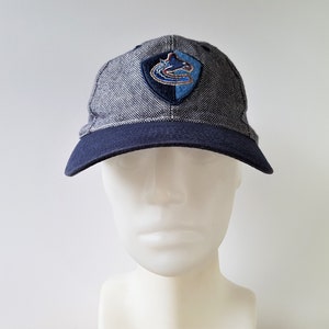 May include: A gray and navy blue baseball cap with a curved brim. The cap features a team logo patch on the front. The patch is a shield shape with a blue and green logo. The cap is displayed on a white mannequin head.