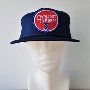May include: A navy blue and white trucker hat with a curved brim. The hat features a circular patch with the text "CARLING O'KEEFE" in white lettering, a red background, and a blue border. A shield graphic is in the center.