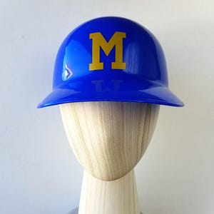 Op de afbeelding: Een glanzende blauwe honkbalhelm met een gele "M" op de voorkant. De helm wordt getoond op een houten hoofdvorm. De helm is een sportartikel.