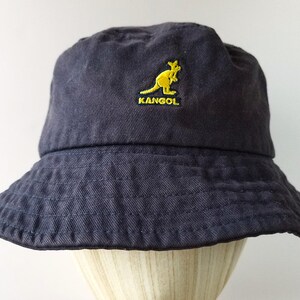K&ouml;nnte beinhalten: Ein marineblauer Fischerhut mit gelbem K&auml;nguru-Logo und dem Wort "KANGOL" auf der Vorderseite gestickt. Der Hut hat eine breite Krempe und ist aus einem weichen, strukturierten Stoff gefertigt.