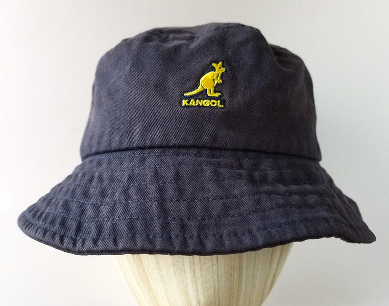 K&ouml;nnte beinhalten: Ein marineblauer Fischerhut mit gelbem K&auml;nguru-Logo und dem Wort "KANGOL" auf der Vorderseite gestickt. Der Hut hat eine breite Krempe und ist aus einem weichen, strukturierten Stoff gefertigt.