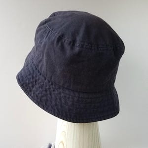 K&ouml;nnte beinhalten: Ein dunkelmarineblauer Fischerhut. Der Hut hat eine abgerundete Krone und eine nach unten geneigte Krempe. Der Hut ist aus einem strukturierten Stoff gefertigt und wird auf einem Holzst&auml;nder vor wei&szlig;em Hintergrund pr&auml;sentiert.