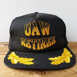 Könnte beinhalten: Schwarze Baseballkappe mit goldfarben gesticktem Schriftzug "UAW RETIREE". Die Kappe hat goldfarbene Blattdetails auf dem Schirm. Ein klassisches Accessoire für Rentner.
