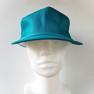 May include: Teal-colored hat with a structured crown and a small brim. The hat features a decorative rope trim around the base of the crown. The interior of the brim is white, with a green edge. The hat is displayed on a white mannequin head.