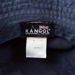 K&ouml;nnte beinhalten: Marineblauer Fischerhut mit einem schwarzen Etikett mit dem Markennamen "KANGOL", dem Text "BORN BRITISH EST. 1938" und der britischen Flagge. Ein wei&szlig;es Etikett lautet "100% COTTON".