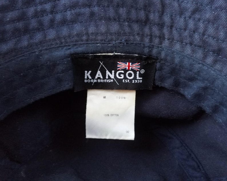 K&ouml;nnte beinhalten: Marineblauer Fischerhut mit einem schwarzen Etikett mit dem Markennamen "KANGOL", dem Text "BORN BRITISH EST. 1938" und der britischen Flagge. Ein wei&szlig;es Etikett lautet "100% COTTON".