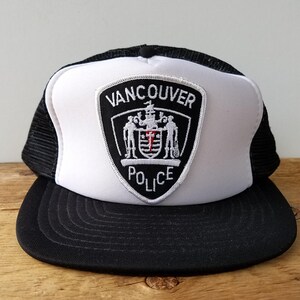Peut inclure: Casquette de camionneur noire et blanche avec une visière noire et un dos en filet. Le panneau avant est blanc avec un écusson "Vancouver Police" et un blason avec un bouclier, des figures et du texte.