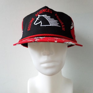 Peut inclure: Casquette de baseball noire et rouge avec un logo de cheval blanc et le texte "Equine Sciences Program".
