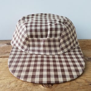 May include: A brown and beige checked cap with a flat peak. The cap features a five-panel design and is made from woven fabric. The pattern is a classic gingham check, suitable for casual wear.