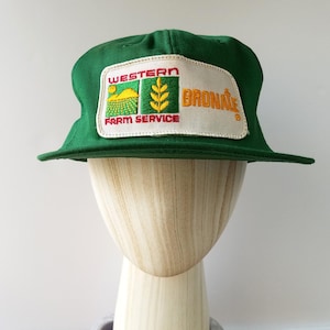 May include: Green baseball cap with a rectangular patch. The patch displays the text "WESTERN FARM SERVICE" and "BRONATE". The design includes yellow and green elements, such as a sun, fields, and wheat.