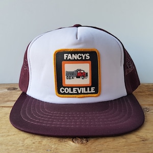 May include: A burgundy and white trucker hat with a square patch featuring a red truck and the words "Fancys Coleville". The hat has a mesh back and a curved brim.