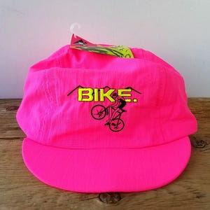 以下が含まれることがあります： 鮮やかなピンク色のサイクリングキャップ。つばはカーブしています。「BIKE」の文字は黄色いブロック体で黒い縁取りがあり、自転車に乗ったサイクリストのグラフィックが描かれています。タグがキャップに付いています。