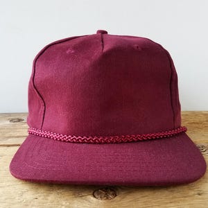 May include: A burgundy baseball cap with a flat brim. The cap features a matching burgundy rope detail along the brim. The cap is made of a solid, textured material.