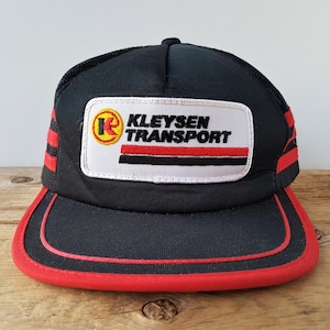 May include: Black trucker hat with red accents and a red brim. The hat features a white patch with the text "KLEYSEN TRANSPORT" in black letters, with a red and black stripe design. The hat has a mesh back.