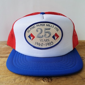 Op de afbeelding: Een witte en blauwe truckercap met een rood mesh-achterpaneel. De pet heeft een patch met de tekst "Island Paper Mills Ltd. 25 Years 1960-1985".