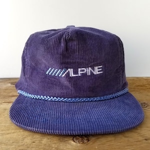 May include: A corduroy baseball cap in shades of purple and blue. The cap features the word "ALPINE" embroidered in white, with a blue and white rope detail around the brim. The hat has a classic, structured design.