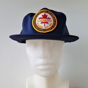 Op de afbeelding: Een blauwe en rode baseballpet met een rond embleem waarop staat "Canadian Association of Industrial Mechanical & Allied Workers - CAIMAW - Founded June 1954".
