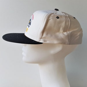 Op de afbeelding: Een wit en zwarte baseballcap met een platte klep. De cap heeft een kleurrijk geborduurd ontwerp op de voorkant en zwarte knopen aan de bovenkant. De klep is zwart en de cap wordt getoond op een mannequinhoofd.