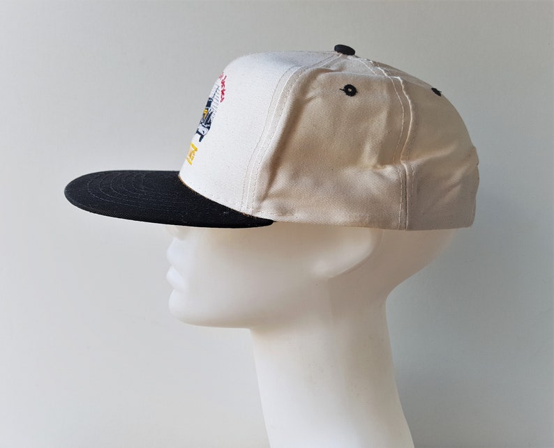 Op de afbeelding: Een wit en zwarte baseballcap met een platte klep. De cap heeft een kleurrijk geborduurd ontwerp op de voorkant en zwarte knopen aan de bovenkant. De klep is zwart en de cap wordt getoond op een mannequinhoofd.