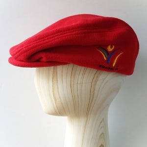May include: A red fleece newsboy cap with a curved brim. The cap features an embroidered emblem with colorful abstract shapes and the text "WINNIPEG '99". The hat is displayed on a wooden mannequin head.