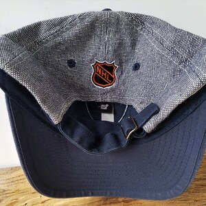 May include: A navy blue baseball cap with a textured gray top and a curved brim. The cap features the NHL logo in orange and black. The adjustable strap is navy blue with a brass-colored buckle.