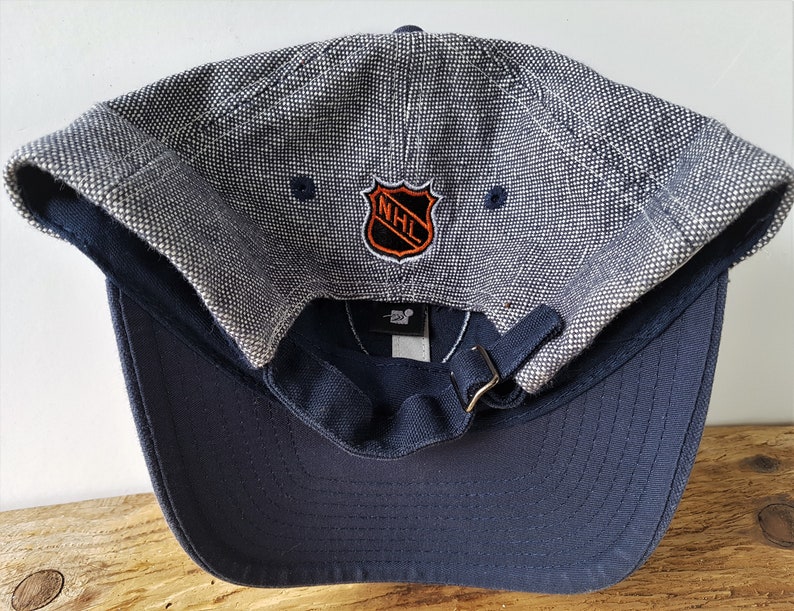 May include: A navy blue baseball cap with a textured gray top and a curved brim. The cap features the NHL logo in orange and black. The adjustable strap is navy blue with a brass-colored buckle.