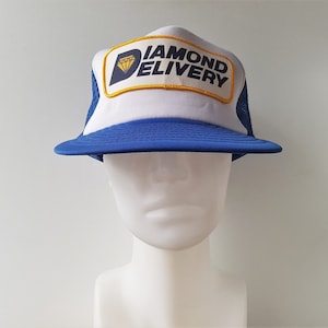 Puede incluir: Un gorro de camionero azul y blanco con un parche amarillo y azul que dice "Diamond Delivery" y un gráfico de diamante.