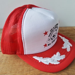 May include: Red and white trucker hat with a mesh back. The front panel is white with a printed design and text that reads "12th Annual Winter Tribal Dakota Council All-Star 1985". The brim is red with white embroidered leaf designs.