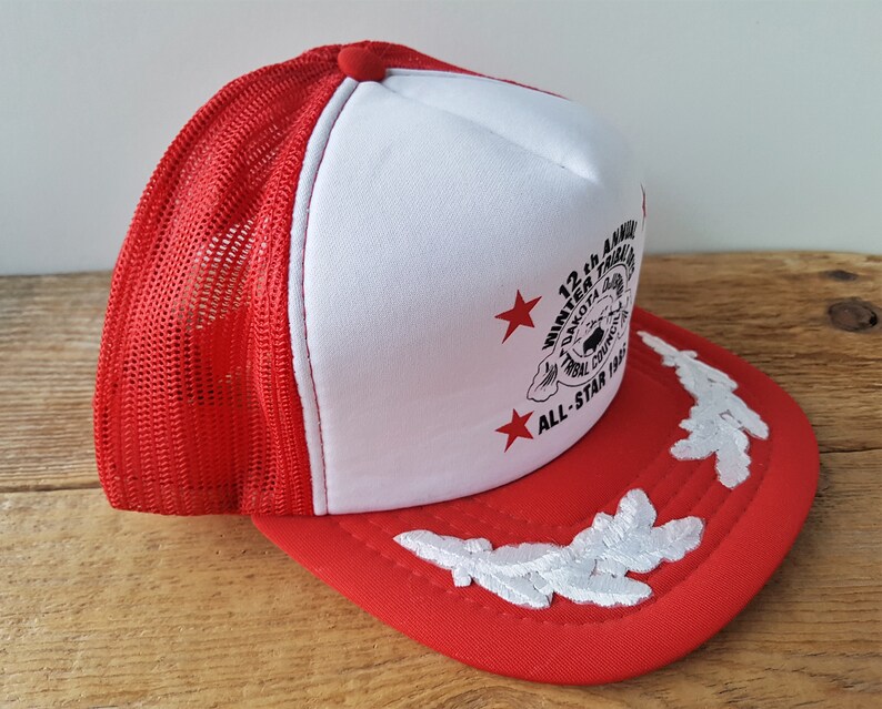 May include: Red and white trucker hat with a mesh back. The front panel is white with a printed design and text that reads "12th Annual Winter Tribal Dakota Council All-Star 1985". The brim is red with white embroidered leaf designs.