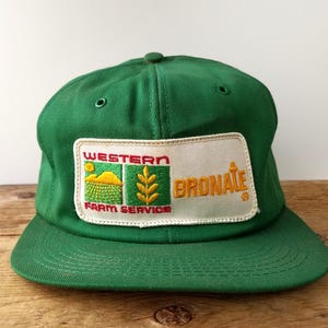 May include: Green baseball cap with a flat peak. The cap has a rectangular patch with the words "WESTERN FARM SERVICE" and "BRONATE" in red and gold lettering. The patch also includes a graphic of a field and wheat.