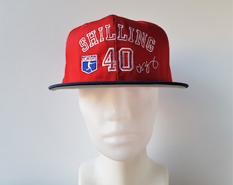 May include: Red baseball cap with a white embroidered logo that says "Shilling 40" and a blue and white logo that says "Players". The cap has a black brim and a white signature.
