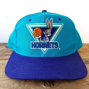May include: A teal and purple Hornets baseball cap. The cap features a Bugs Bunny graphic holding a basketball inside a white triangle. The word "HORNETS" is embroidered below the graphic.