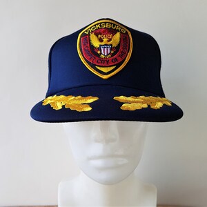 May include: A navy blue baseball cap with a gold embroidered patch that says "Vicksburg Police" and a gold eagle with a red shield. The cap has two gold embroidered oak leaves on the front.