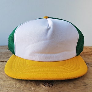 Può includere: Cappellino da camionista con pannello frontale bianco, retro in rete verde e visiera gialla. Il cappello ha un bottone giallo sulla parte superiore. Il cappello è su una superficie di legno.