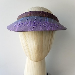 Op de afbeelding: Een paarse zonnevizier met de tekst "Oyster Bay Golf Club" geborduurd op de voorkant. De zonnevizier heeft een donkerbruine en paarse band. De zonnevizier wordt getoond op een houten mannequinhoofd.