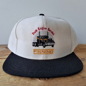 Op de afbeelding: Een cr&egrave;mekleurige baseballcap met een zwarte klep. De pet heeft een geborduurd ontwerp van een zwart-gele vrachtwagen met de tekst "Truck Engine Service" en "FINNING" in rood en goud.