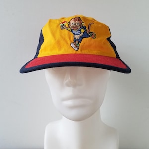 Peut inclure: Une casquette de baseball jaune, rouge et bleue avec un singe de dessin animé portant une salopette et tenant une fleur. Le singe est brodé sur le devant de la casquette.