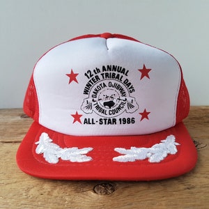 May include: Red and white trucker hat with a mesh back. The front features the text "12th Annual Winter Tribal Days Dakota Ojibway Tribal Council All-Star 1986" and red star graphics. White embroidered leaf designs adorn the brim.
