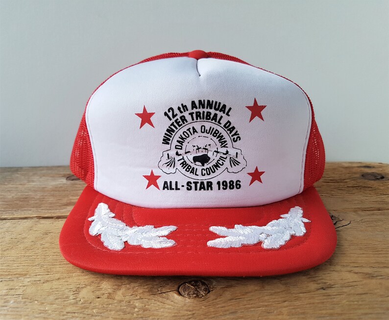 May include: Red and white trucker hat with a mesh back. The front features the text "12th Annual Winter Tribal Days Dakota Ojibway Tribal Council All-Star 1986" and red star graphics. White embroidered leaf designs adorn the brim.