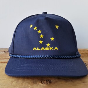 Puede incluir: Una gorra de béisbol azul marino con ribete de cuerda amarillo y el diseño de la bandera del estado de Alaska que presenta un contorno amarillo de la constelación de la Osa Mayor y la palabra "ALASKA" en letras amarillas.