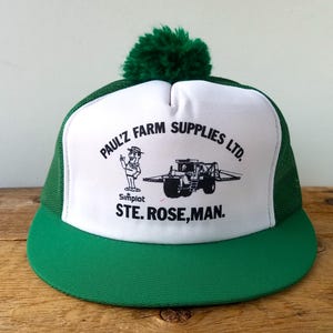Può includere: Un cappellino da camionista verde e bianco con un pompon verde. Il pannello frontale mostra il testo "PAUL'Z FARM SUPPLIES LTD." e un'illustrazione di un agricoltore e un trattore. È visibile anche il testo "STE. ROSE, MAN."