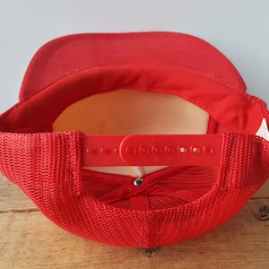 May include: A red trucker hat with a mesh back and a plastic snap closure. The hat has a curved brim and a red adjustable strap. The interior of the hat is a light yellow color.