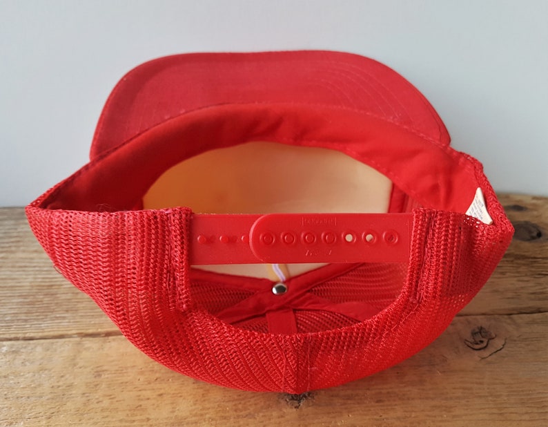 May include: A red trucker hat with a mesh back and a plastic snap closure. The hat has a curved brim and a red adjustable strap. The interior of the hat is a light yellow color.