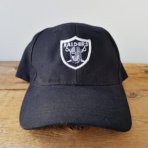 May include: Black baseball cap with a curved brim. The cap features a white and gray embroidered Raiders logo on the front. The logo is a shield shape with the word "RAIDERS" at the top and a graphic of a football player.