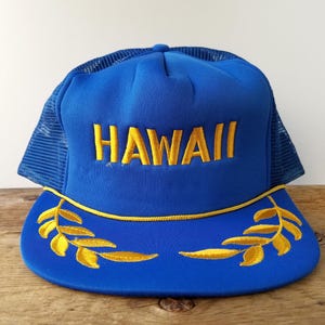 Könnte beinhalten: Blaue Trucker-Mütze mit dem Wort "HAWAII" in Gelb auf der Vorderseite gestickt. Die Mütze hat ein gelbes, gesticktes Blattdesign auf dem Schirm und eine Mesh-Rückseite.