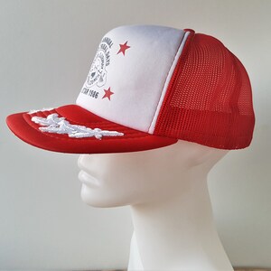 May include: A red and white trucker hat with a mesh back. The hat features a red brim with white embroidered detailing and a white front panel with text and star graphics. The text reads "ANNUAL FESTIVAL DAYS" and "STAR 1986".