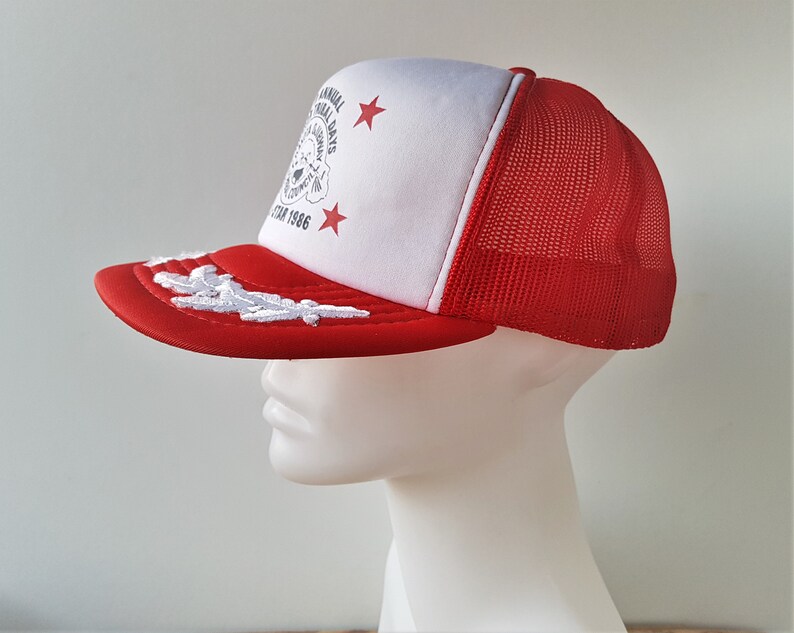 May include: A red and white trucker hat with a mesh back. The hat features a red brim with white embroidered detailing and a white front panel with text and star graphics. The text reads "ANNUAL FESTIVAL DAYS" and "STAR 1986".